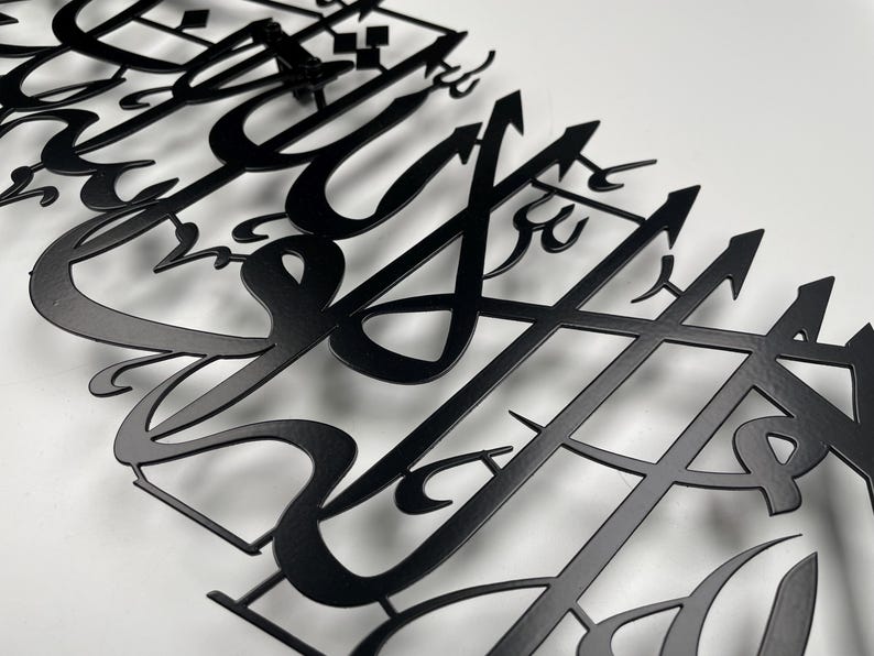 May include: Close-up of a black metal wall art piece featuring intricate Arabic calligraphy. The artwork is cut from metal and mounted on a white surface, showcasing the detailed design and elegant script. The piece is a decorative item.