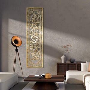 Könnte beinhalten: Ein goldfarbenes, rechteckiges Wandkunstwerk mit aufwendiger arabischer Kalligraphie. Das Kunstwerk wird vertikal präsentiert und hat einen dekorativen Rahmen. Es ist an einer strukturierten, grauen Wand angebracht und verleiht der Raumdekoration Eleganz.