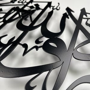 May include: Close-up of black metal Arabic calligraphy art. The intricate design features flowing, stylized letters with a matte finish. The artwork is set against a white background, highlighting the detailed cut-out shapes and forms.