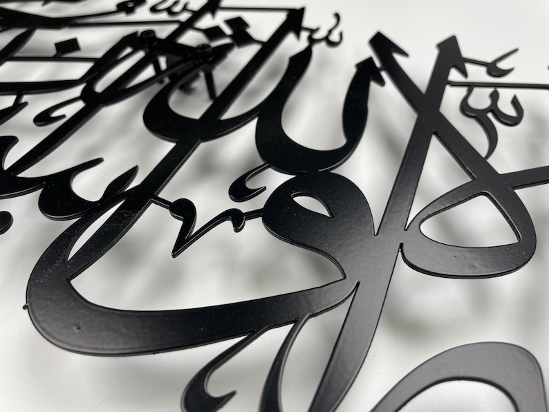 May include: Close-up of black metal Arabic calligraphy art. The intricate design features flowing, stylized letters with a matte finish. The artwork is set against a white background, highlighting the detailed cut-out shapes and forms.