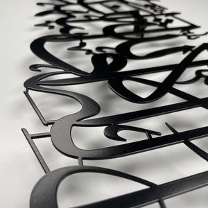 May include: Black metal wall art featuring stylized Arabic calligraphy. The artwork is cut from metal and mounted on a white surface. The design has a modern aesthetic, with flowing lines and intricate details, suitable for home decor.