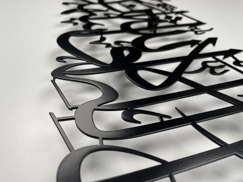 May include: Black metal wall art featuring stylized Arabic calligraphy. The artwork is cut from metal and mounted on a white surface. The design has a modern aesthetic, with flowing lines and intricate details, suitable for home decor.