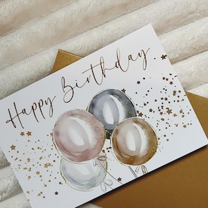 May include: A white card with the text "Happy Birthday." in gold script. The card features a watercolor illustration of four balloons in shades of pink, gray, and brown with gold glitter accents. The background is scattered with gold stars.