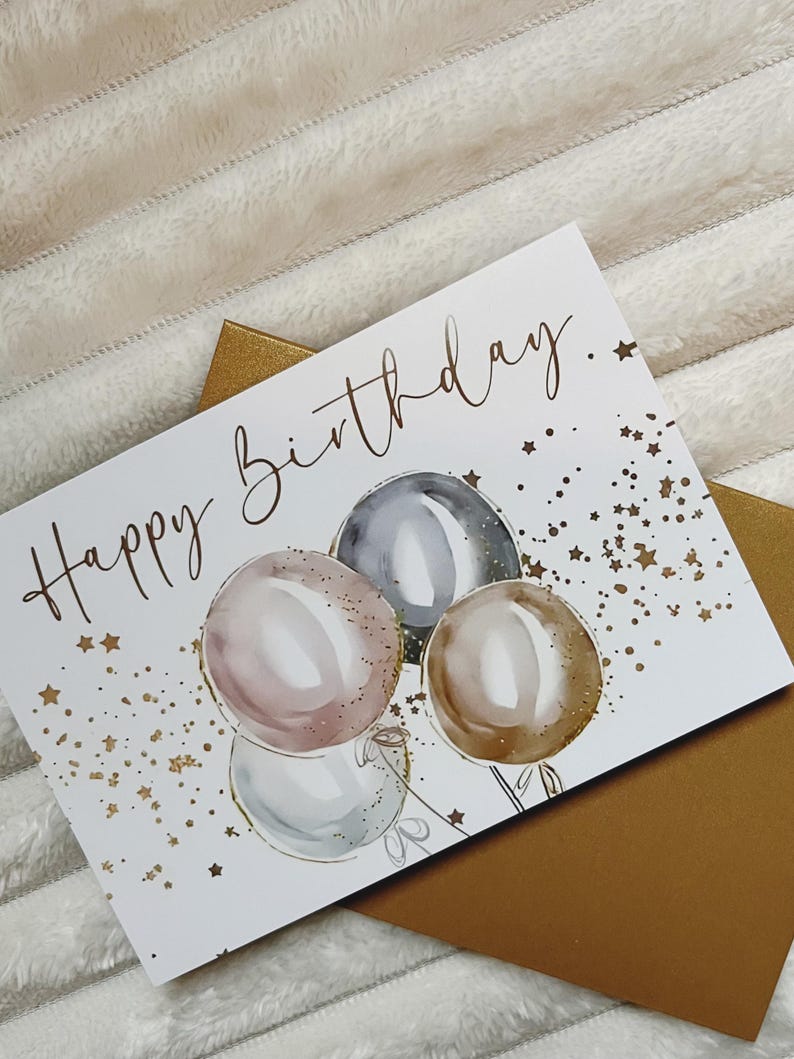 May include: A white card with the text "Happy Birthday." in gold script. The card features a watercolor illustration of four balloons in shades of pink, gray, and brown with gold glitter accents. The background is scattered with gold stars.