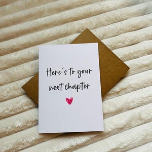May include: A white card with black text that reads "Here's to your next chapter" with a pink heart below the text. The card is on a brown envelope.
