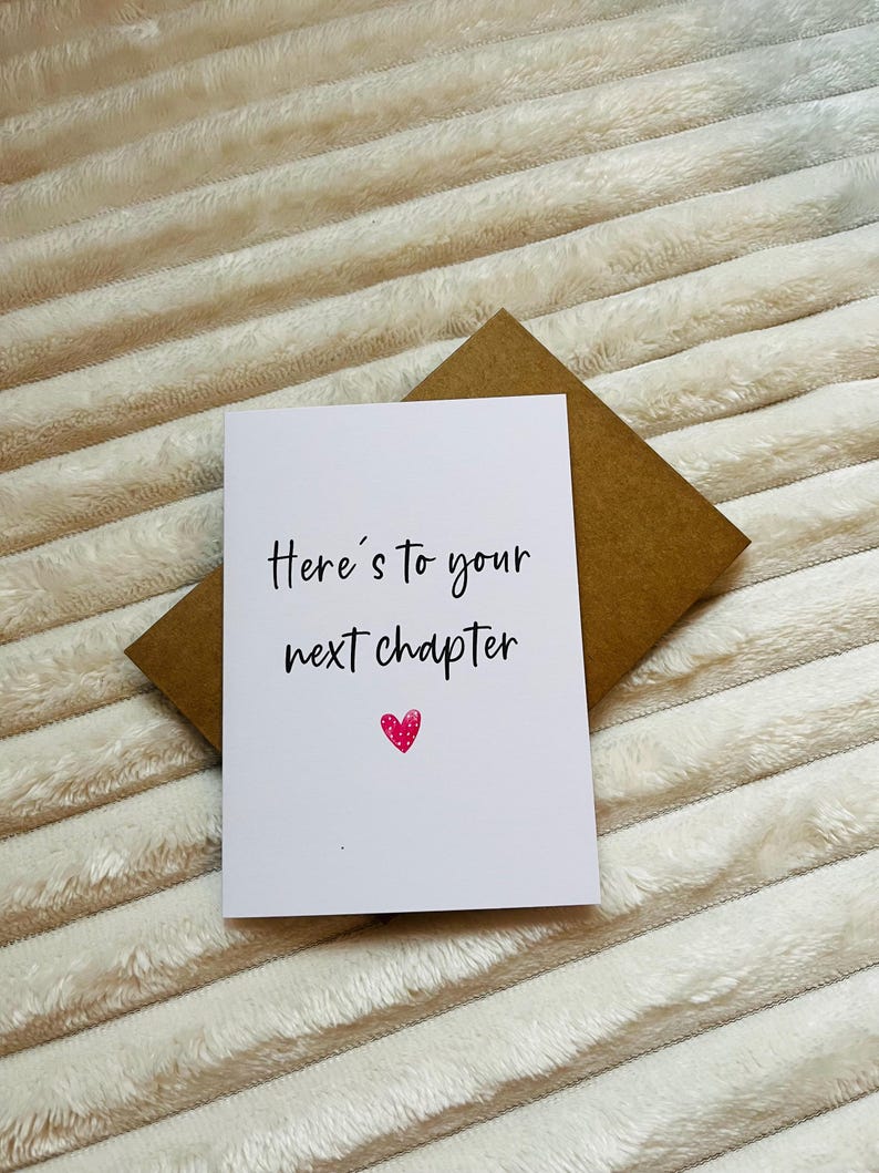 May include: A white card with black text that reads "Here's to your next chapter" with a pink heart below the text. The card is on a brown envelope.