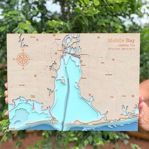 May include: A layered wooden bathymetric map of Mobile Bay, Alabama, USA. The map features light wood landmasses with engraved roads and city names, and water depths represented by stacked blue and turquoise layers. A compass rose is on the left.