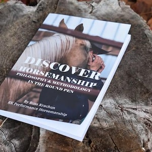 May include: A book titled "Discover Horsemanship" is displayed on a weathered tree stump. The cover shows a horse's head and a person. The title and subtitle are in white text. The book is about philosophy and methodology.