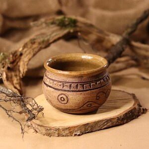 Op de afbeelding: Een kleine, bruine keramische pot met een getextureerd oppervlak en ingewikkelde ontwerpen. De pot staat op een houten schijf.