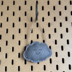 May include: A gray felt cloud ornament with a smiling face and embroidered details. The cloud is suspended by a natural twine string. The ornament is hanging against a brown pegboard background.