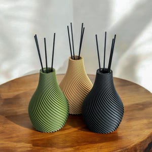 Può includere: Tre bottiglie decorative per diffusore con canne nere. Le bottiglie hanno la forma di una pera con un motivo a spirale. Le bottiglie sono verdi, beige e nere.