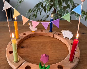 May include: A circular wooden candle ring featuring four lit candles in yellow, light green, blue, and red. The ring is decorated with a small wooden goose, a sheep, a pink flower, and a string of colourful felt pennant flags between two poles.