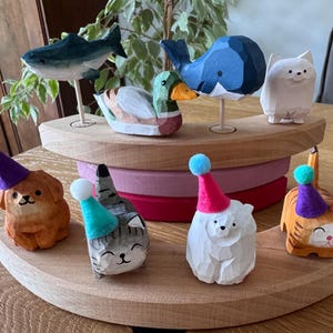 Könnte beinhalten: Handgeschnitzte Holzfiguren von Tieren, darunter ein Hai, ein Wal, eine Ente, eine Katze, ein Hund, ein Eisbär und ein Tiger, jeweils mit einer Partyhütchen. Die Figuren sind auf einem mehrstufigen Holzständer ausgestellt, der die detaillierte Handwerkskunst zeigt.