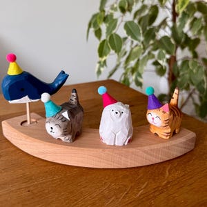 Könnte beinhalten: Ein geschwungener Holzsockel trägt vier kleine geschnitzte Tierfiguren: einen dunkelblauen Wal auf einem Stab, eine graue getigerte Katze, einen weißen Bären und eine orangefarbene getigerte Katze. Jedes Tier trägt einen kleinen, bunten Partyhut mit Bommel.