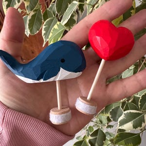 Könnte beinhalten: Handgeschnitzte Holzfiguren, eine Wal und ein Herz. Der Wal ist blau und weiß, das Herz rot. Beide Figuren haben Holzstäbe als Beine und sind an kleinen, runden Sockeln mit weißem Filzbesatz befestigt.