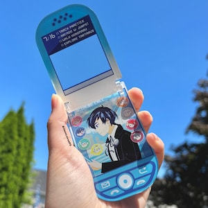 May include: A blue and clear acrylic flip phone featuring a cartoon character illustration. The phone shows the text "Incoming Call" and a list of options. The phone is held against a blue sky.
