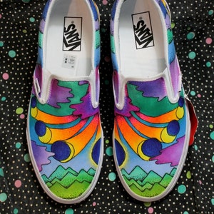 May include: A pair of white canvas slip-on shoes with a colorful, abstract design. The design features a rainbow, clouds, and mountains. The shoes have the Vans logo on the side.