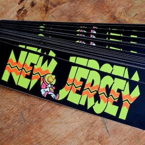 May include: A stack of black and green stickers with the words "NEW JERSEY" in a yellow and orange zig-zag pattern. A cartoon figure wearing a sombrero is in the lower left corner.