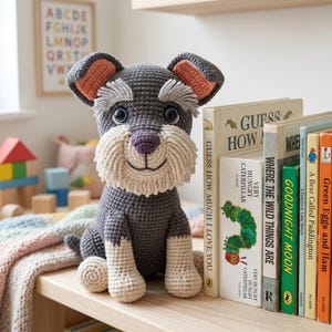 May include: A dark grey crocheted dog plush toy with a cream-coloured muzzle, eyebrows, and paws. The toy features large dark eyes, a purple nose, and pink-lined ears. It is seated upright with a textured yarn finish, displaying a friendly expression.