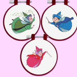 May include: Three circular embroidery hoops containing cross-stitch patterns of three fairy characters. One fairy wears a green dress, one wears a blue dress, and one wears a pink dress. Each fairy holds a wand and is depicted against white fabric.