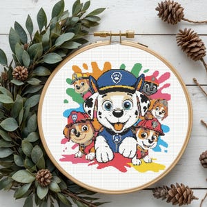 May include: A cross-stitch embroidery hoop featuring the Paw Patrol characters. The design includes the main characters with colorful paint splatters in the background. The hoop is set against a white wooden background with greenery and pinecones.