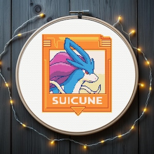 Könnte beinhalten: Kreuzstich-Stickerei von Suicune, einem blauen und lila Pokémon, eingerahmt in einem orangefarbenen Rahmen. Das Wort "SUICUNE" wird darunter angezeigt. Die Stickerei befindet sich in einem Holzrahmen mit Lichterketten.