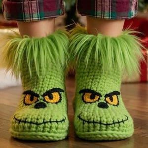 Op de afbeelding: Een paar felgroene gehaakte pantoffels met een pluizige rand van imitatiebont aan de bovenkant. De pantoffels zijn versierd met geborduurde gele ogen, zwarte wenkbrauwen en een gestikte zwarte mond, wat een karakteristieke gezichtsuitdrukking geeft.