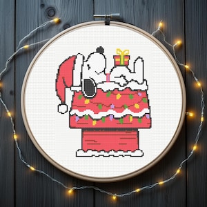 May include: A cross-stitch embroidery of Snoopy in a Santa hat, resting on his Christmas-decorated doghouse. The doghouse is red with colourful lights and a gift on top. The embroidery is in a wooden hoop, with fairy lights around it.