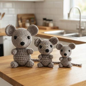May include: Three grey crocheted mice of varying sizes arranged in a row. Each mouse features a rounded body, large circular ears, black bead eyes, and a small black nose. The figures are crafted with a textured yarn stitch and have long tails.