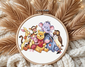 May include: A cross-stitch embroidery featuring Winnie the Pooh and friends, including Piglet, Tigger, Eeyore, and Owl. The characters are in a wooden embroidery hoop, with a white background. The artwork is surrounded by dried palm leaves and a white knitted blanket.