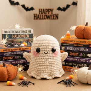 May include: A cream-coloured crocheted ghost plushie with black button eyes and pink cheeks, positioned among stacks of books titled Where the Wild Things Are Halloween, Ghosts in the Library, and Spooky ABCs, alongside fabric pumpkins and plastic spiders.