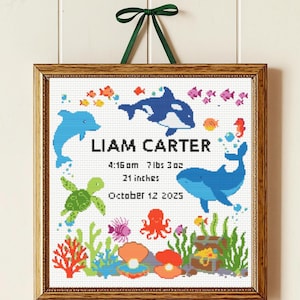 Puede incluir: Cuadro de punto de cruz enmarcado, con tema submarino, que muestra un delfín, una orca y una ballena. Se muestra el nombre "Liam Carter", con detalles de nacimiento: 3,3 kg, 53,3 cm, y 12 de octubre de 2025. Colgado con una cinta verde.