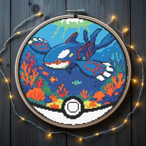 May include: Cross-stitch art of a Kyogre, a blue and red sea creature, swimming in a blue ocean with orange coral and small fish. The design is framed in a wooden hoop, resembling a Poke Ball. Fairy lights surround the hoop.