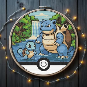 May include: Cross-stitch embroidery featuring two blue turtle-like creatures in a natural setting, framed in a wooden hoop. The design includes a waterfall and greenery, with a white and black Poké Ball design at the bottom.