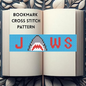 May include: A bookmark cross stitch pattern featuring the word "JAWS" with a shark in the middle. The shark is gray with red teeth and a white mouth. The letters are red and the background is blue.