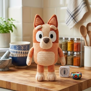 Peut inclure: Une peluche au crochet en forme de chien faite de fil orange et crème, avec de larges taches blanches autour des yeux, un nez marron et des oreilles pointues. Le jouet a un corps arrondi, de petits bras et deux jambes, posé debout.