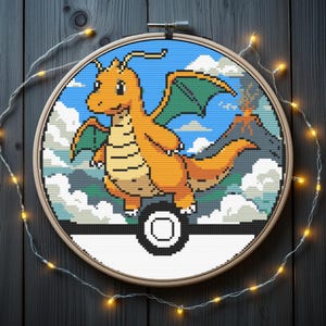 Puede incluir: Un bastidor de bordado circular con un patrón de punto de cruz de una criatura naranja similar a un dragón con alas verdes. El fondo muestra un cielo azul, nubes blancas y un volcán. El diseño está enmarcado por una esfera blanca y negra.