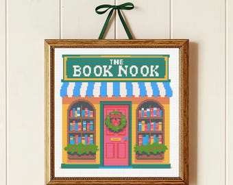 May include: Cross-stitch artwork of a bookstore facade. The sign reads "THE BOOK NOOK" above a blue and white striped awning. The building has a pink door with a wreath and bookshelves on either side, framed in a wooden frame and hung with a green ribbon.
