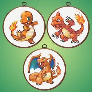 May include: Three cross-stitch hoops displaying Pokémon characters Charmander, Charmeleon, and Charizard. The characters are in orange, yellow, and blue, set against a white background. The hoops have wooden frames and metal hanging loops.