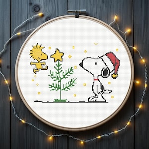Könnte beinhalten: Kreuzstich-Stickerei mit Snoopy mit Weihnachtsmütze, der einen Weihnachtsbaum mit Stern betrachtet. Woodstock fliegt in der Nähe. Das Design befindet sich auf weißem Stoff, umgeben von einem Holzrahmen und Lichterketten.