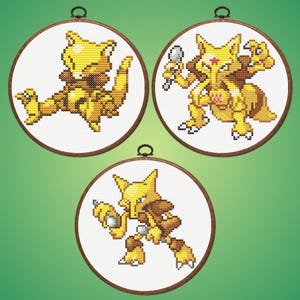 Könnte beinhalten: Drei runde Stickrahmen zeigen jeweils einen anderen gelb-braunen, pixeligen Pokémon-Charakter. Die Charaktere sind vor einem weißen Hintergrund platziert, mit einem dunkelbraunen Holzreifen und einer kleinen Metallschlaufe zum Aufhängen.