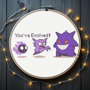 Puede incluir: Un bastidor de bordado circular de madera con tela blanca y un diseño de punto de cruz de tres criaturas moradas en progresión. El texto "You've Evolved!" está bordado en negro sobre las dos primeras figuras de la secuencia.