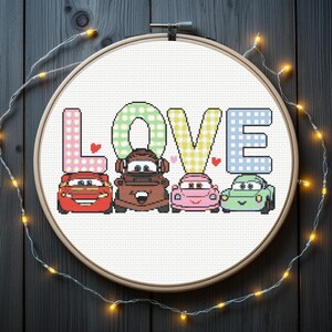 以下が含まれることがあります： 木製のフレームに入ったクロスステッチ刺繍で、4台の漫画の車の上の「LOVE」という言葉が特徴です。文字はピンク、緑、黄色、青で模様が描かれています。小さな赤いハートが文字の近くに配置されています。フレームはライトのストリングで囲まれています。