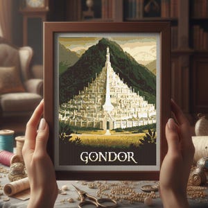 May include: A framed cross-stitch depicting Gondor, a city from a fantasy setting, with a mountain backdrop. The artwork is in a brown wooden frame. The word "GONDOR" is stitched at the bottom. The image is held by hands.