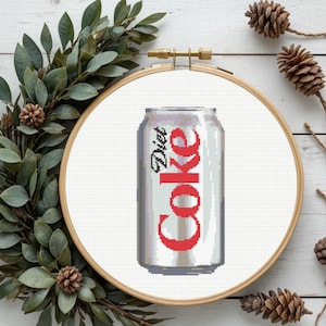Pode incluir: Um design de ponto cruz de uma lata de Diet Coke num bastidor de bordado de madeira. A lata é prateada com as palavras "Diet Coke" em vermelho. O bastidor é rodeado por folhas verdes e pinhas.