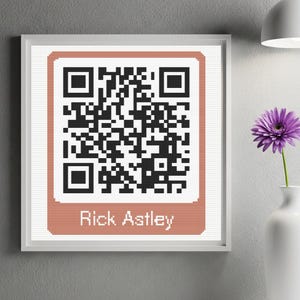 May include: Framed artwork featuring a pixelated QR code in black and white, set against a peach-colored background. The text "Rick Astley" is below the code. A white vase with a purple flower is to the right.