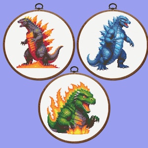 May include: Three round embroidery hoops each featuring a pixelated Godzilla design. One Godzilla is black with orange flames, another is blue, and the third is green with orange flames. Each design is set against a white background.