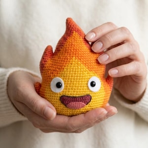Puede incluir: Un pequeño peluche de ganchillo con forma de llama que presenta un degradado de hilo amarillo, naranja y rojo. El personaje tiene dos grandes ojos de fieltro blanco con pupilas negras, una boca de color morado oscuro y una pequeña lengua rosa.