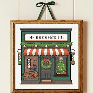 Op de afbeelding: Kruissteekafbeelding van een kapsalon met een rood-wit gestreepte luifel, een kerstkrans en een kerstboom in het raam. Het bord boven de winkel luidt "THE BARBER'S CUT". Het kunstwerk is ingelijst in een houten lijst en heeft een groen lint.