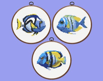 May include: Three framed cross-stitch artworks featuring colorful fish. Each fish is a different species, with blue, yellow, and white hues. The round frames are made of wood and have a metal loop for hanging. The background is a solid light purple.